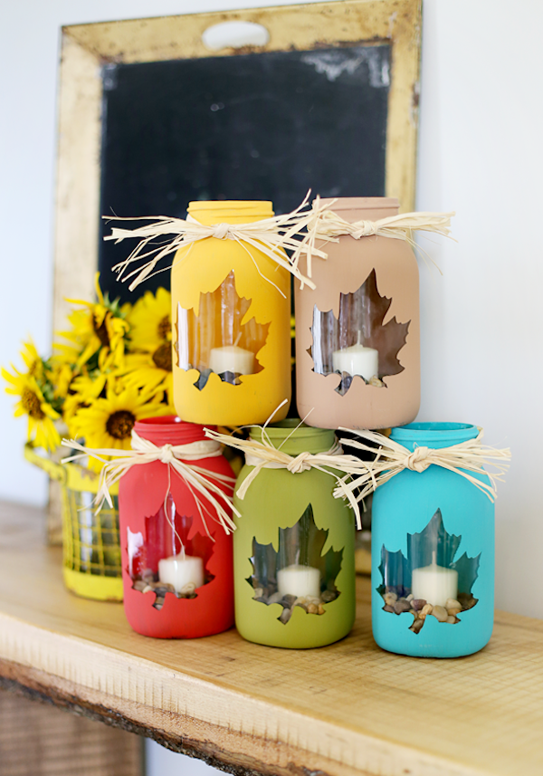 Colorful painted mason jar candle holders with leaf cutouts and raffia bows for DIY Thanksgiving décor.