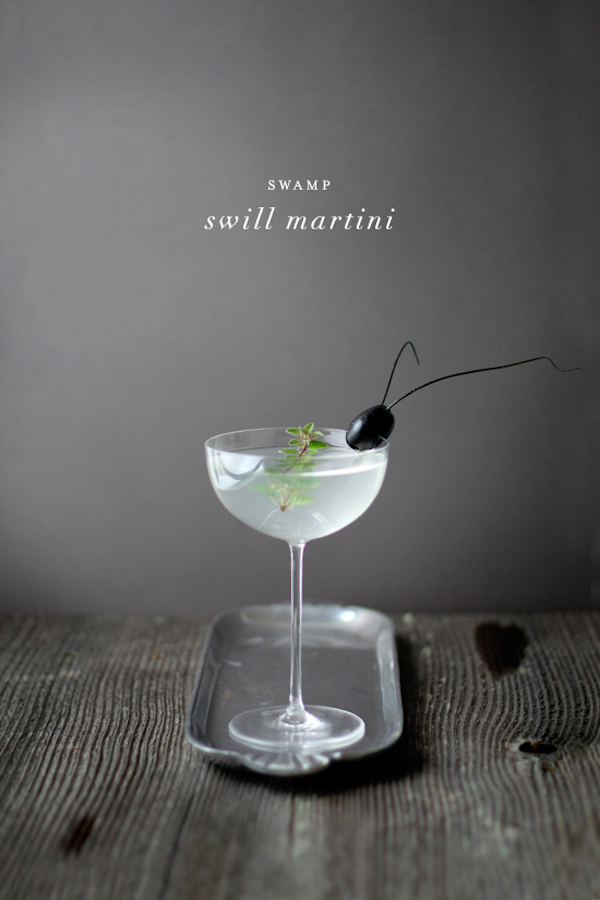Swamp swill martini in a delicate coupe glass, garnished with a sprig of oregano and a whimsical “Palmetto Bug” made from a black olive with chive antennae. The glass sits on a narrow silver tray against a dark gray background, creating a moody, elegant Halloween aesthetic.