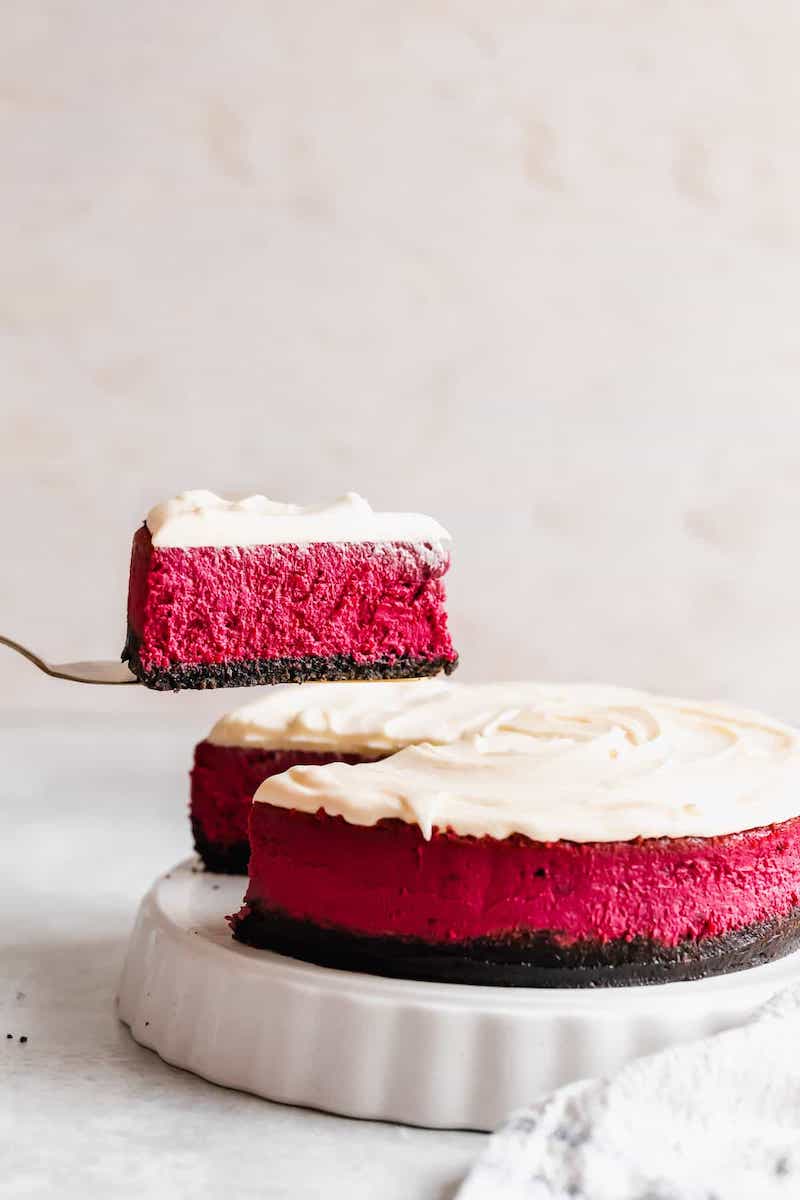 Slice of red velvet cheesecake being lifted from a whole cake on a white stand. The cheesecake has a dark Oreo crust, a vibrant red velvet filling, and a smooth cream cheese frosting layer on top. The background is softly lit, highlighting the rich red color and creamy texture of the dessert.