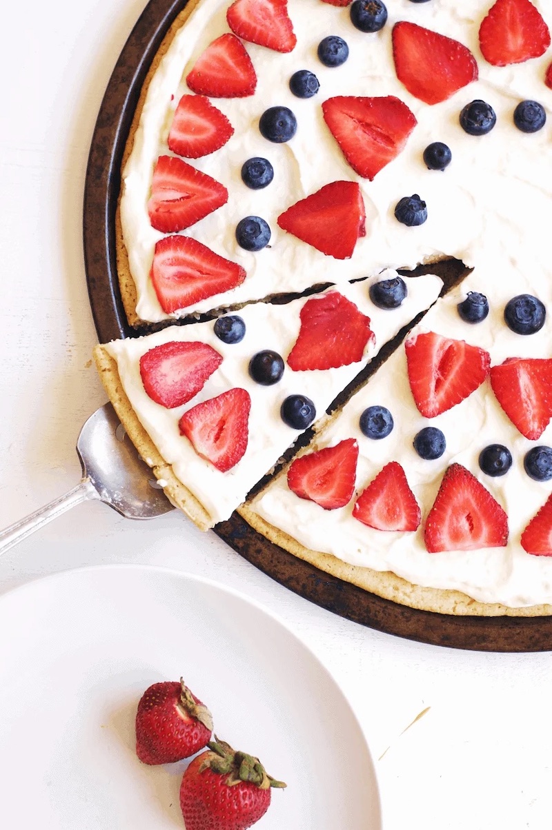 Sugar cookie dessert pizza topped with cream cheese frosting, fresh strawberries, and blueberries for a festive red, white, and blue 4th of July dessert.