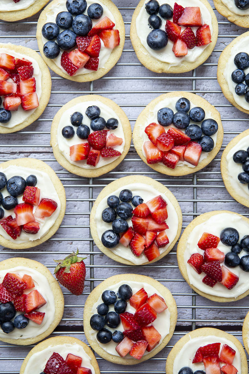 Mini sugar cookie fruit pizzas topped with cream cheese frosting, fresh strawberries, and blueberries for an easy red, white, and blue Fourth of July dessert.