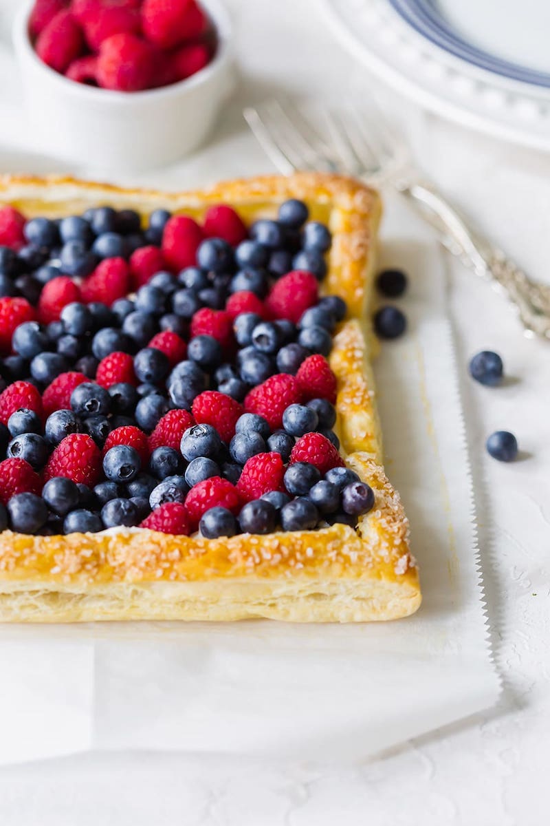 Flaky puff pastry tart topped with a creamy filling, fresh blueberries, and raspberries for an elegant red, white, and berry summer dessert.