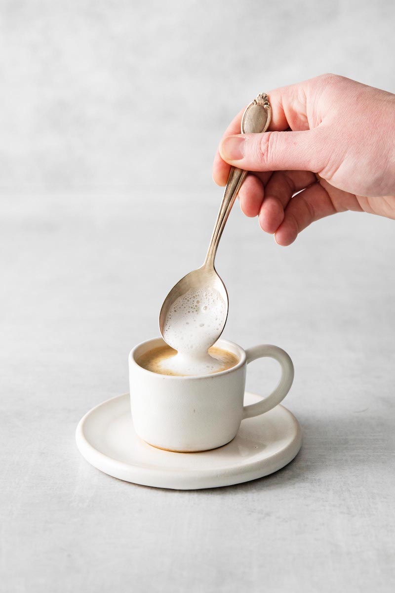 A spoon adds a dollop of creamy milk foam to a freshly made espresso macchiato in a small white espresso cup on a matching saucer.