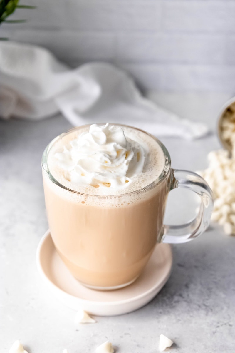 A homemade white chocolate mocha topped with whipped cream in a clear glass mug on a matching saucer.