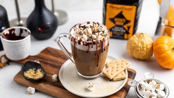 A mug of s'mores witch's brew coffee cocktail with whipped cream on top and chocolate sauce, coffee, and mini pumpkins in the background.