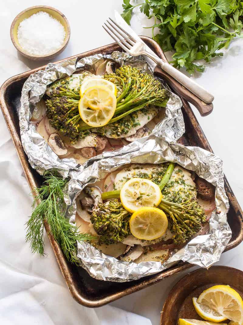 Tilapia foil packet with broccolini, potatoes, mushrooms, red onion, lemon slices, and fresh herbs cooked in aluminum foil.