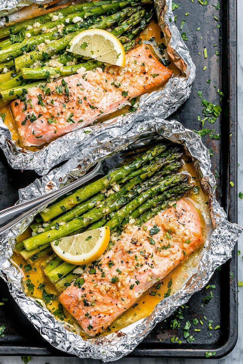 Salmon and asparagus cooked in foil packets with garlic butter, fresh parsley, and lemon wedges on a baking sheet.