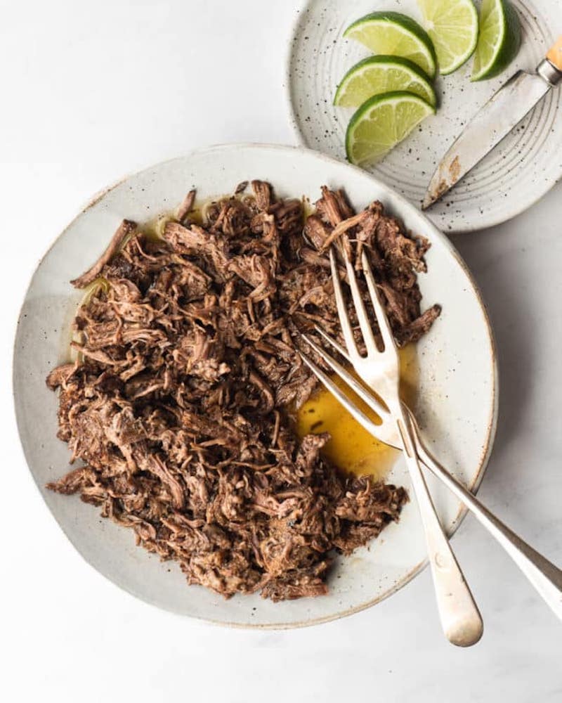 A grey plate filled with shredded barbacoa beef glistening with juices, two forks resting on the side, and a small plate of lime wedges with a knife nearby on a marble surface.