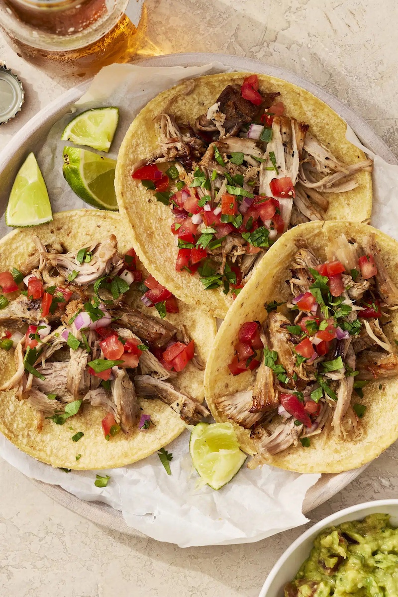 Three corn tortillas filled with shredded slow-cooked pork carnitas topped with fresh pico de gallo made of diced tomatoes, onions, and cilantro, served with lime wedges on a plate beside a bowl of guacamole and a glass of beer.