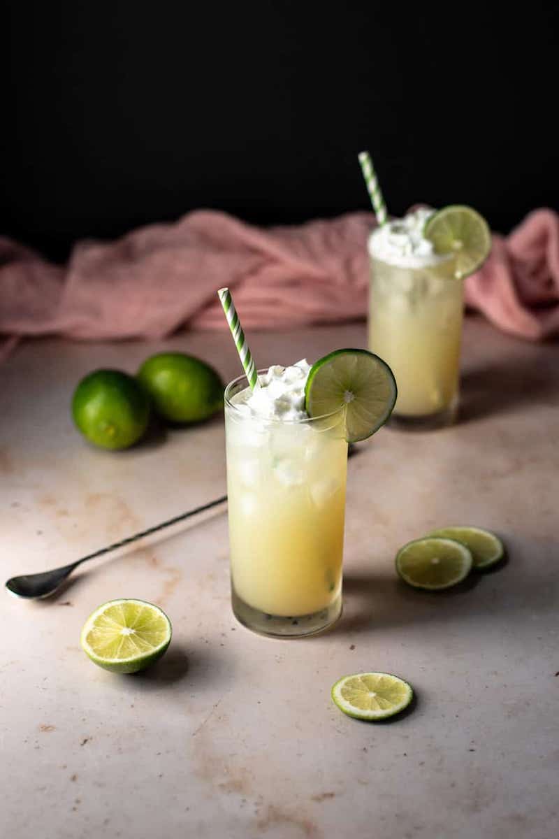 Two tall glasses of key lime pie crush cocktail with straws and lime wedge garnish.
