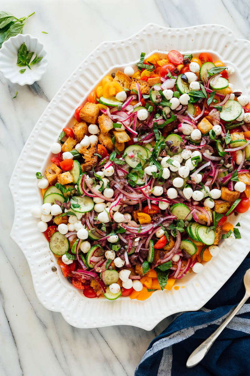 Layered Panzanella Salad with tomatoes, cucumbers, fresh mozzarella, red onion, basil, homemade croutons, and vinaigrette on a white serving platter.