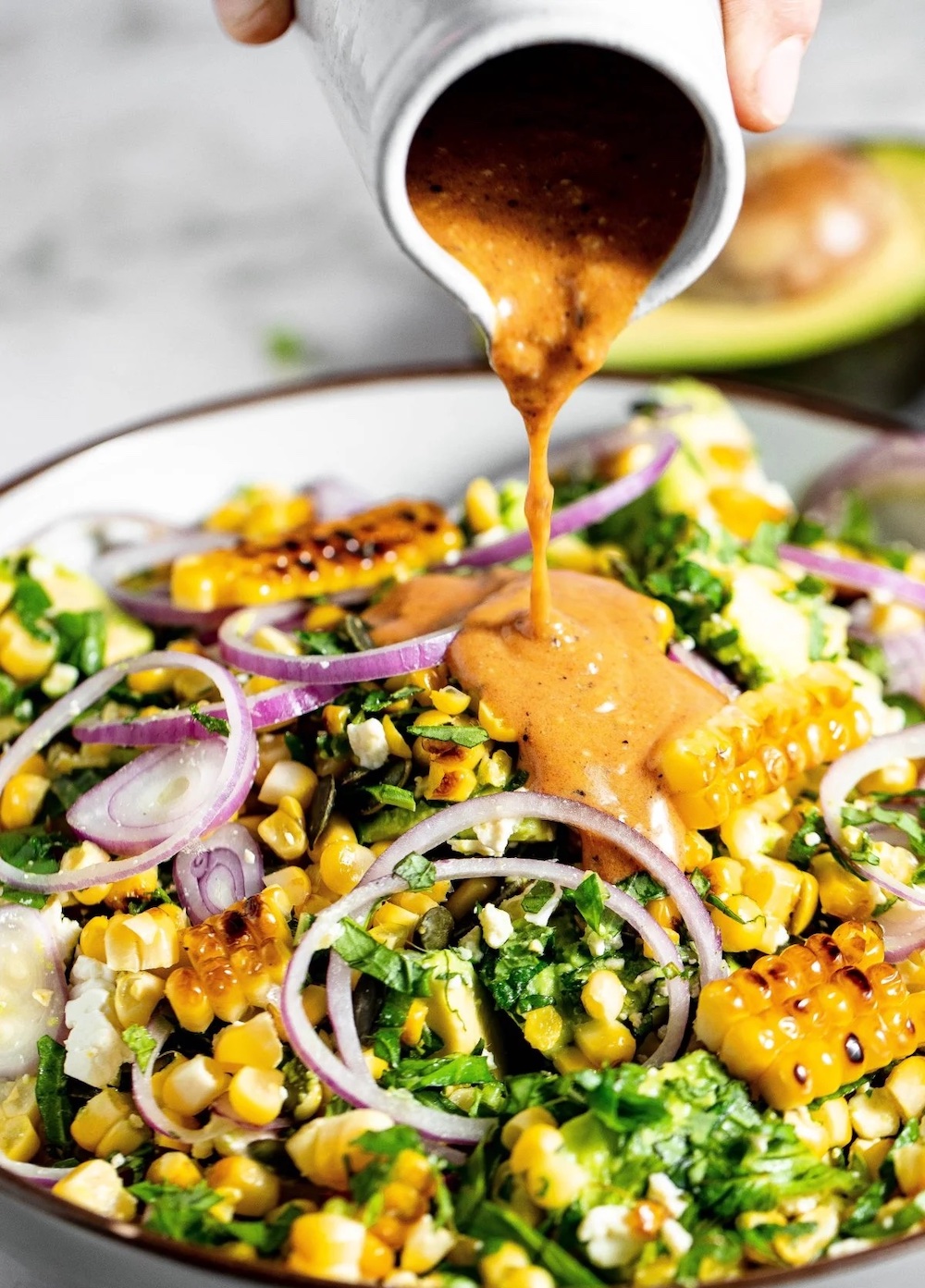 Fresh summer corn salad with grilled corn, avocado, feta, red onion, herbs, and creamy dressing being poured over the top.
