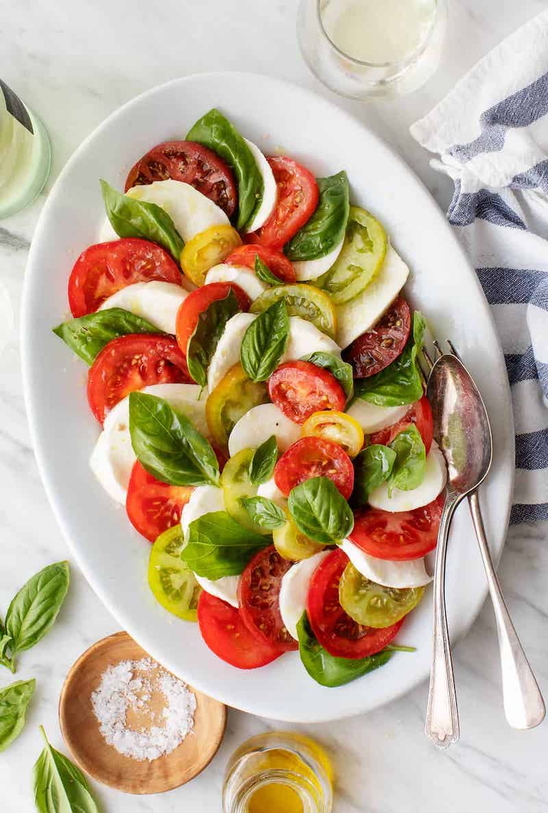 Classic Caprese salad with sliced heirloom tomatoes, fresh mozzarella, basil leaves, olive oil, and balsamic dressing on a white serving platter.
