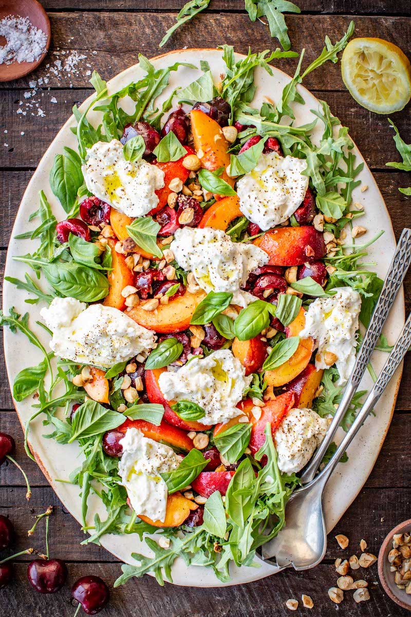 Cherry Peach Burrata Salad with fresh peaches, cherries, creamy burrata, arugula, basil, toasted hazelnuts, and a lemon dressing on a large serving platter.