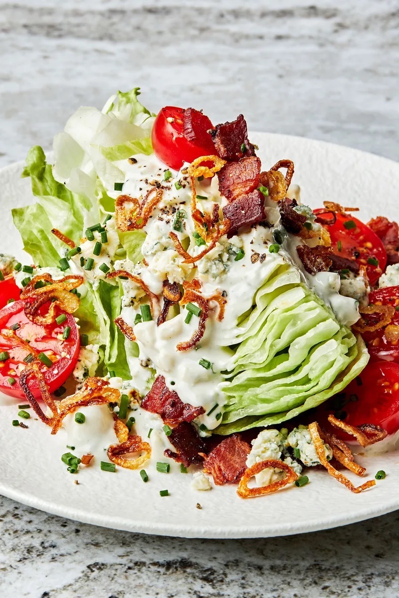 Classic Wedge Salad with iceberg lettuce, blue cheese dressing, crispy bacon, cherry tomatoes, fried onions, and chives on a white plate.