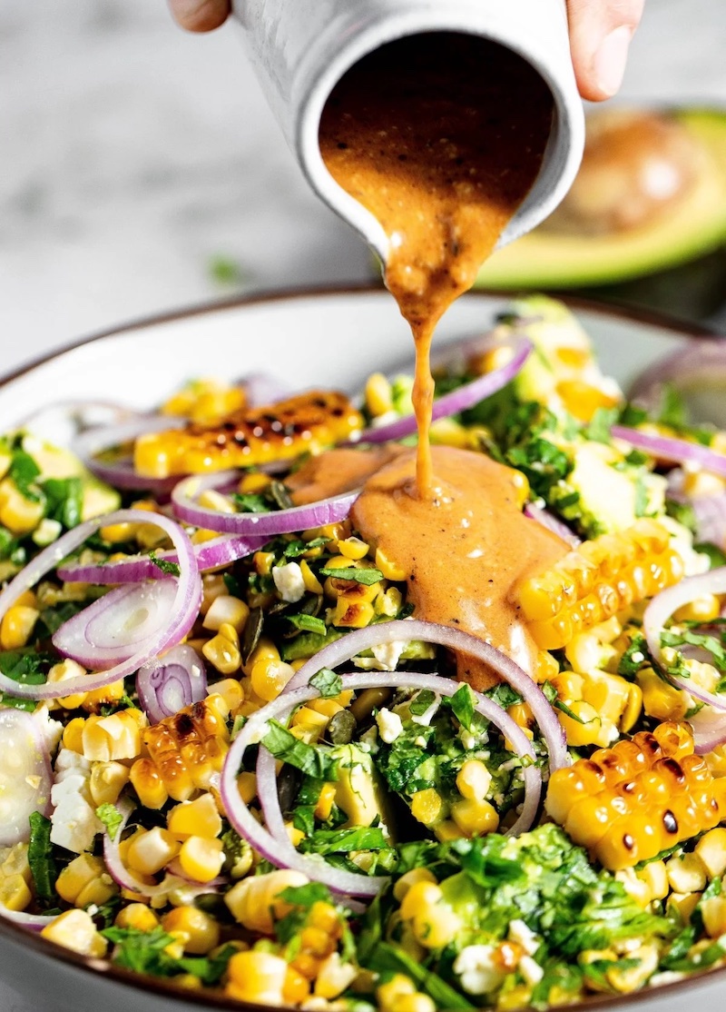 Grilled corn and avocado salad with charred corn, avocado, red onion, fresh herbs, feta cheese, and creamy dressing being poured over the salad.