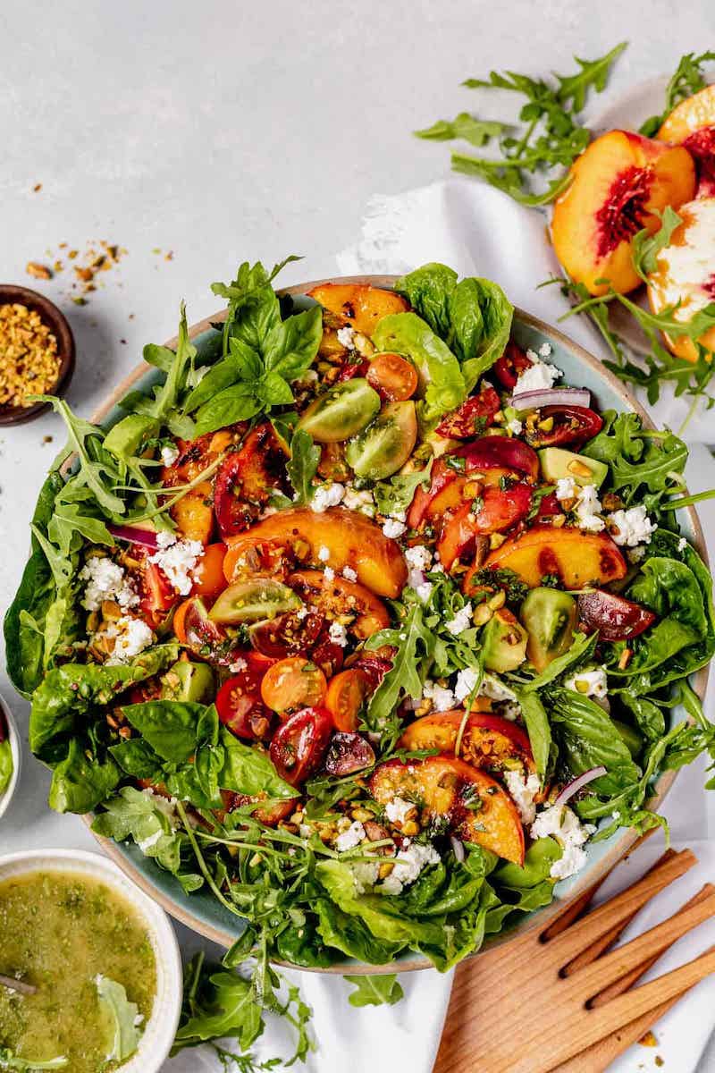 A colorful grilled peach salad made with mixed greens, grilled peach slices, cherry tomatoes, avocado, goat cheese, basil, red onion, and pistachios, tossed with a fresh basil vinaigrette.