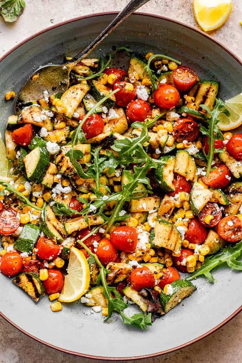 Grilled Zucchini Salad with grilled zucchini, cherry tomatoes, sweet corn, arugula, crumbled feta cheese, and lemon wedges on a large serving platter.