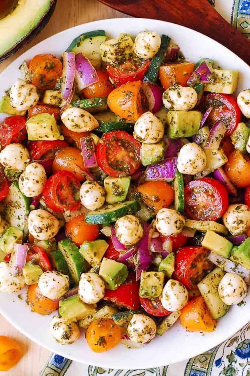 A fresh tomato cucumber avocado salad made with cherry tomatoes, cucumber, avocado, mozzarella pearls, red onion, and basil pesto dressing in a large serving bowl.