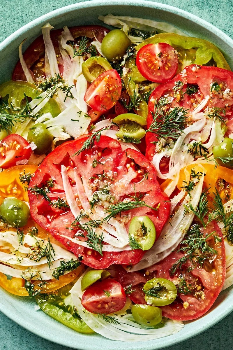 Tomato fennel salad with sliced heirloom tomatoes, shaved fennel, green olives, fresh dill, and white wine vinaigrette in a serving bowl.