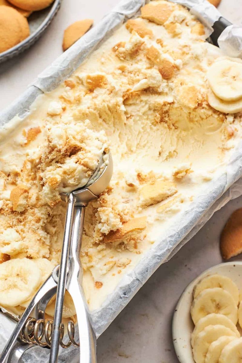 Creamy banana pudding ice cream with sliced bananas and crushed Nilla Wafers in a pie dish with an ice cream scoop.