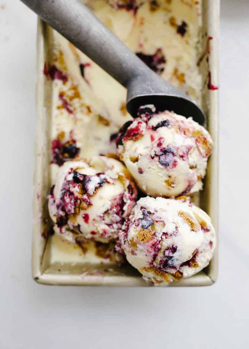 Three scoops of blueberry mascarpone ice cream swirled with blueberry sauce and crunchy Biscoff cookie crumbles in a loaf pan, with a metal ice cream scoop resting on top.