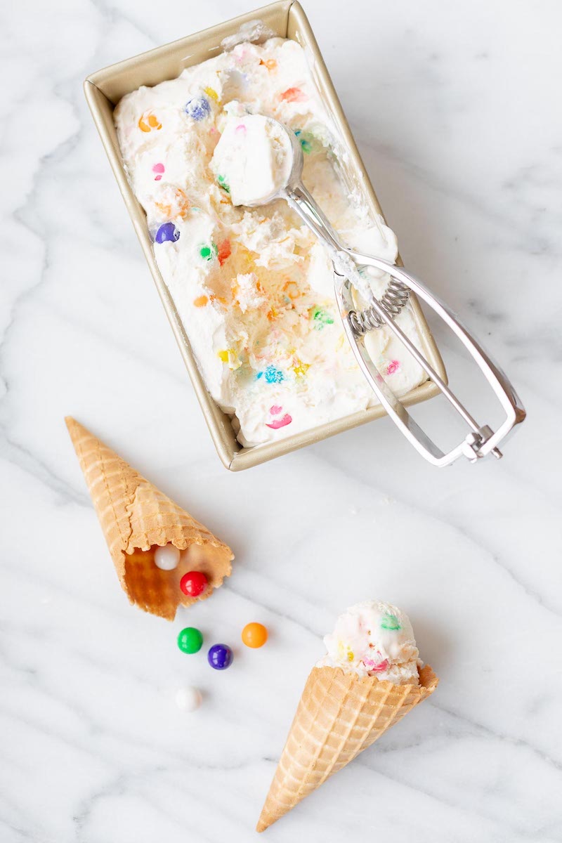 Overhead view of creamy no-churn bubble gum ice cream with colorful bubble gum candy pieces in a loaf pan and served in waffle cones.
