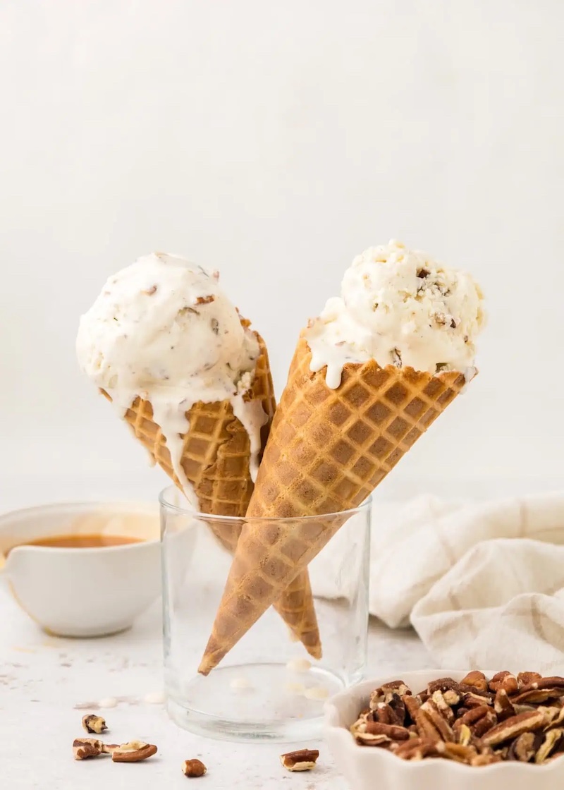 Homemade butter pecan ice cream scooped into waffle cones with toasted pecans and caramel sauce on a white tabletop.