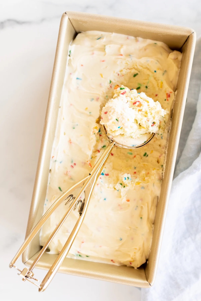 Overhead view of creamy no-churn cake batter ice cream with colorful rainbow sprinkles in a loaf pan and a gold ice cream scoop.