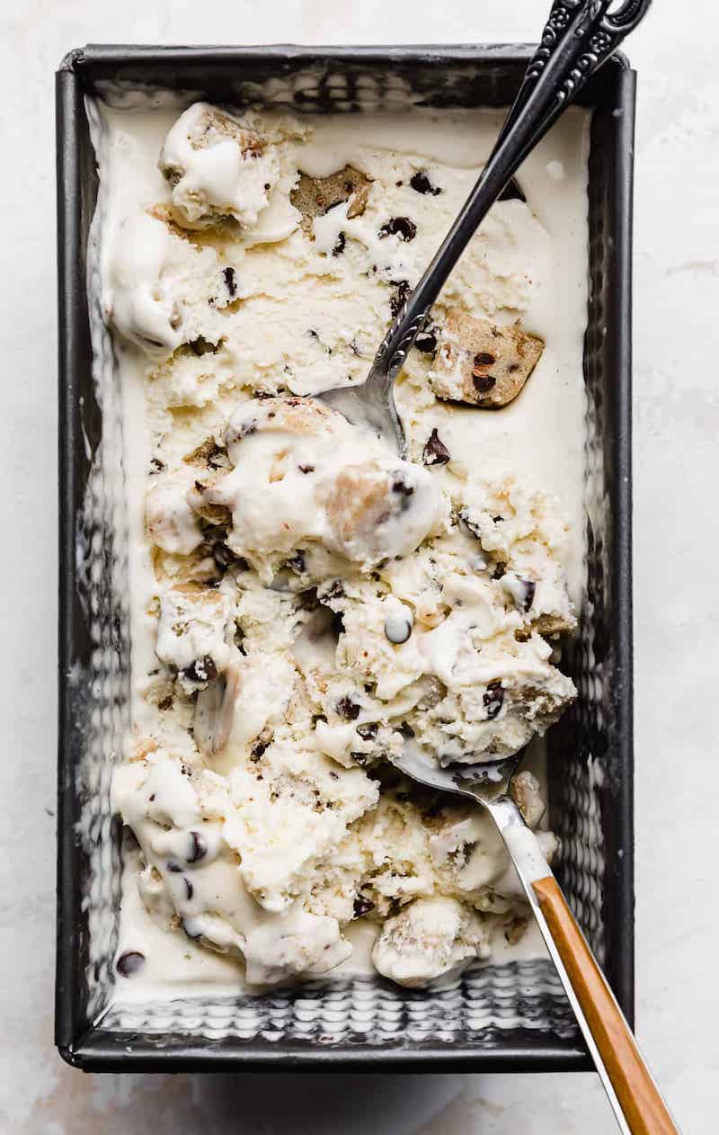 Homemade chocolate chip cookie dough ice cream in a loaf pan with edible cookie dough pieces, mini chocolate chips, and two serving spoons.