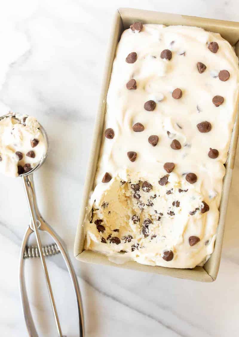 Overhead view of creamy no-churn chocolate chip ice cream in a loaf pan topped with chocolate chips, with an ice cream scoop filled with chocolate chip ice cream beside it.