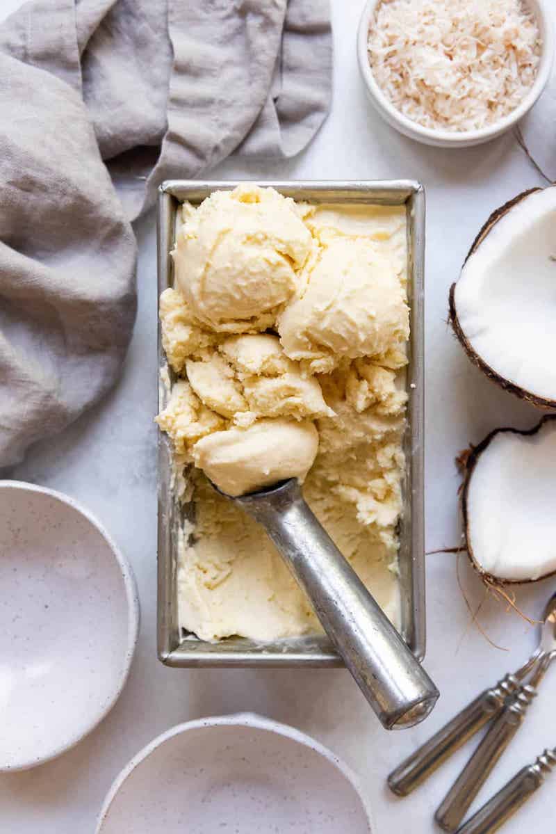 Homemade coconut ice cream in a loaf pan with a scoop, surrounded by fresh coconut halves and toasted coconut flakes.