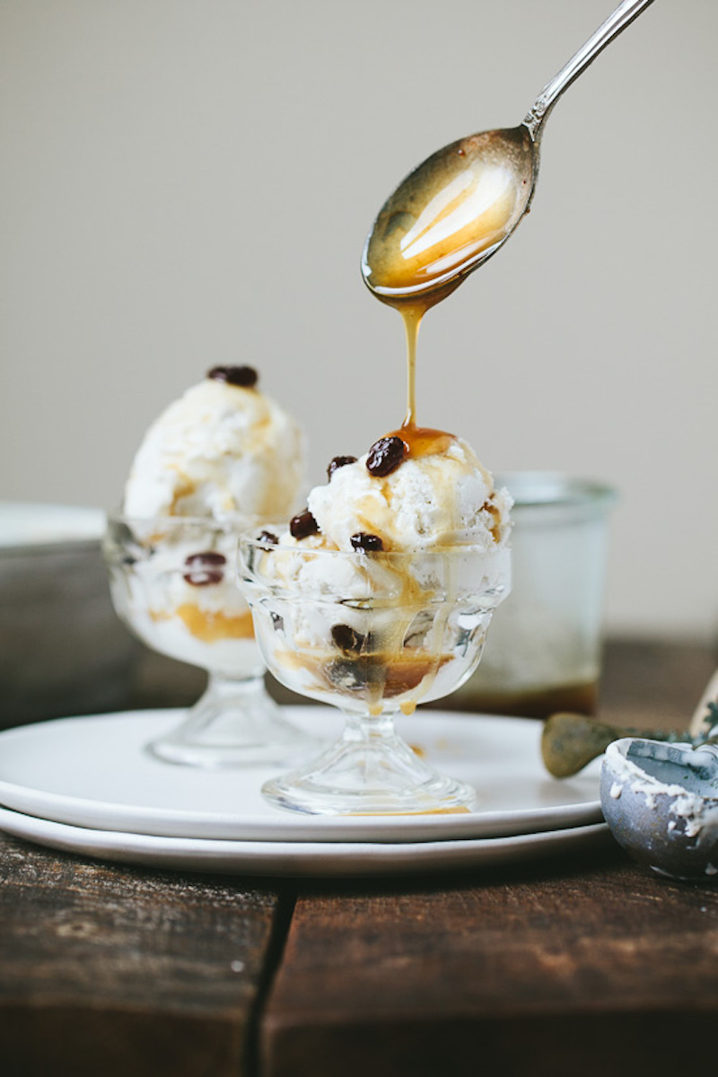 Dairy-free coconut vanilla bean ice cream topped with warm maple rum raisin sauce and raisins, served in glass dessert dishes.