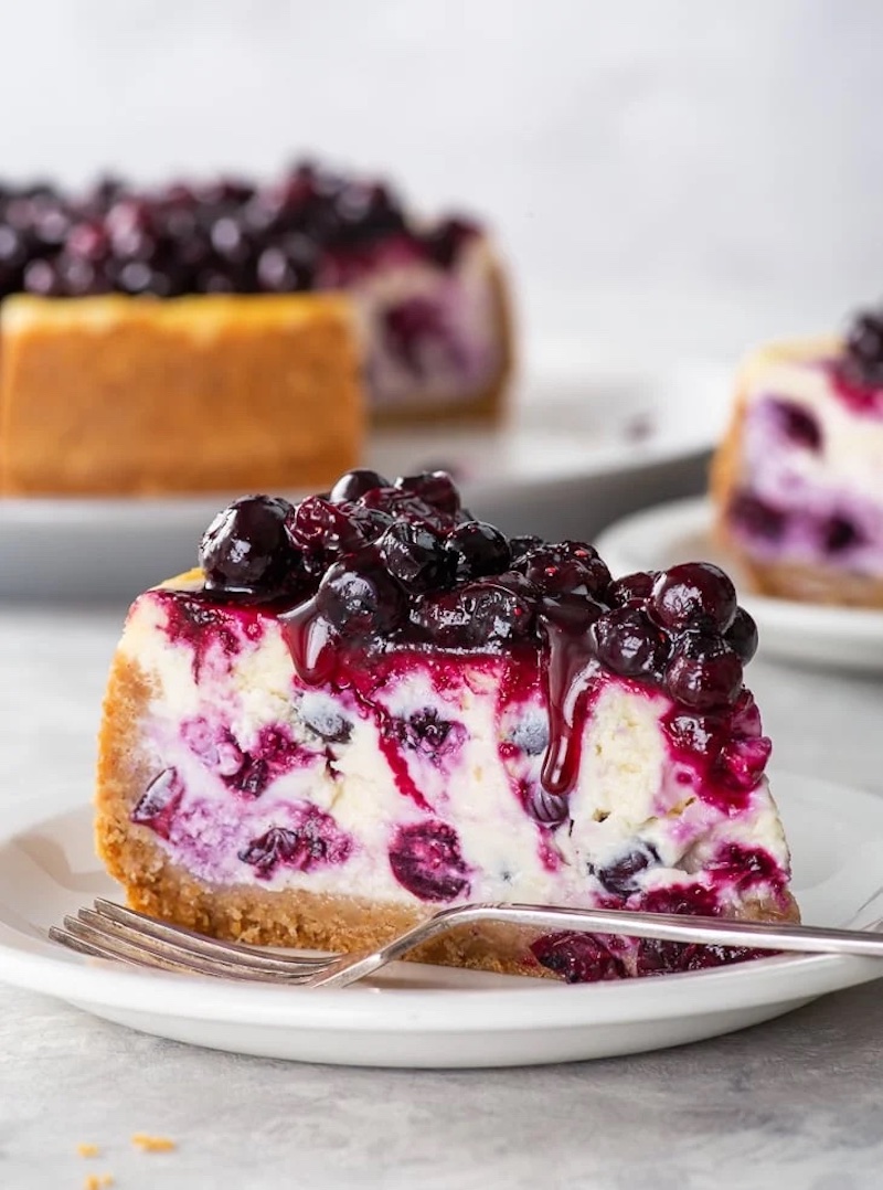 Slice of blueberry cheesecake topped with juicy macerated blueberries and extra blueberry juice dripping down the sides.