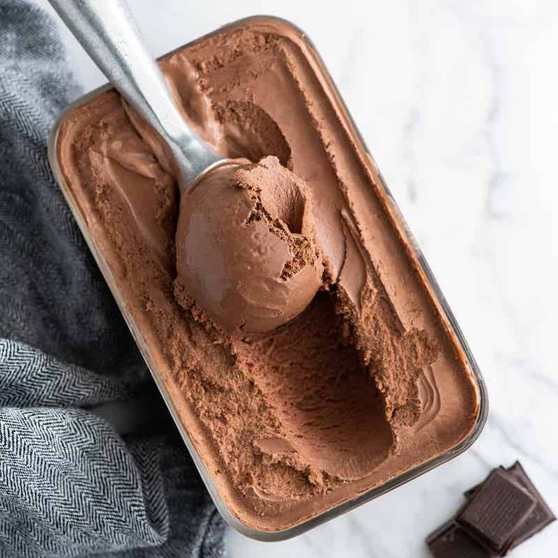 Creamy homemade chocolate ice cream scooped from a loaf pan with pieces of dark chocolate nearby.
