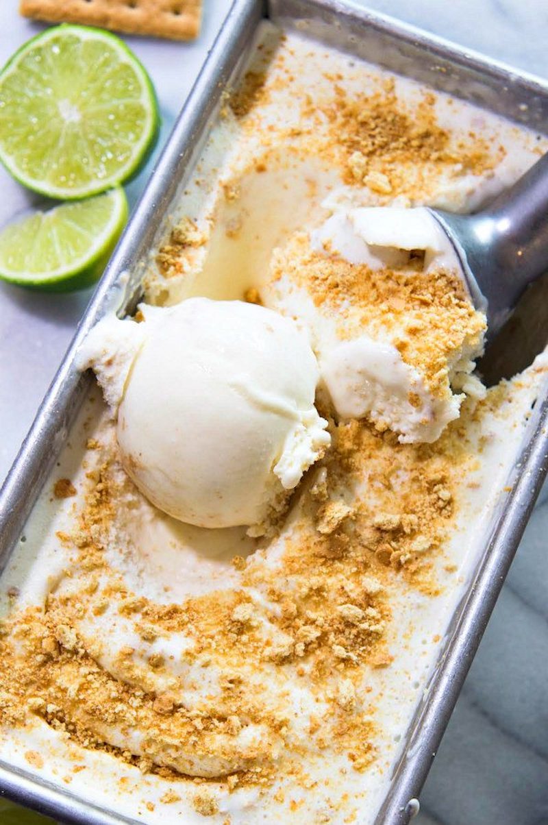 Homemade key lime pie ice cream in a loaf pan topped with graham cracker crumbs, served with fresh lime halves and an ice cream scoop.