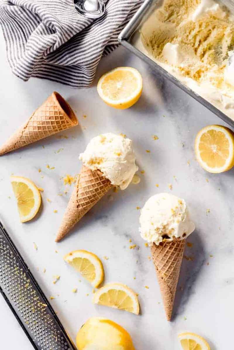 Scoops of homemade fresh lemon ice cream in waffle cones with fresh lemon slices, lemon zest, and a loaf pan of ice cream in the background.