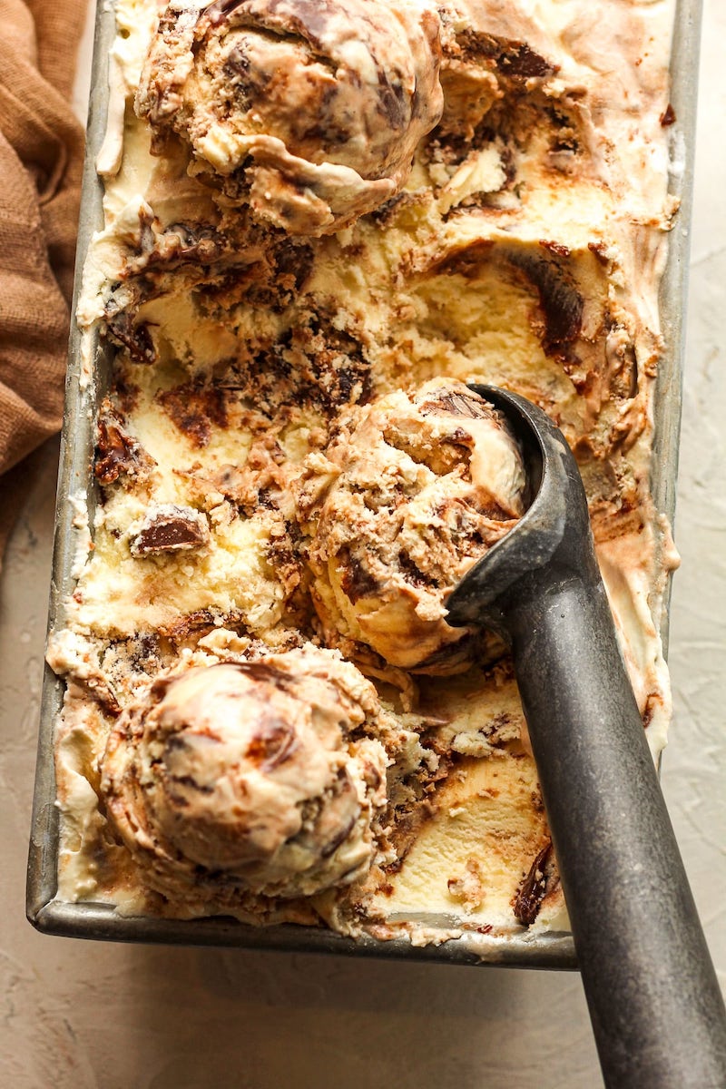 Homemade Moose Tracks ice cream swirled with hot fudge and mini peanut butter cups in a loaf pan with an ice cream scoop.