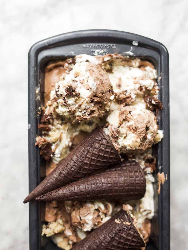 Overhead view of no-churn Half Baked ice cream swirled with chocolate and vanilla ice cream, brownie pieces, and edible cookie dough in a loaf pan with chocolate waffle cones.