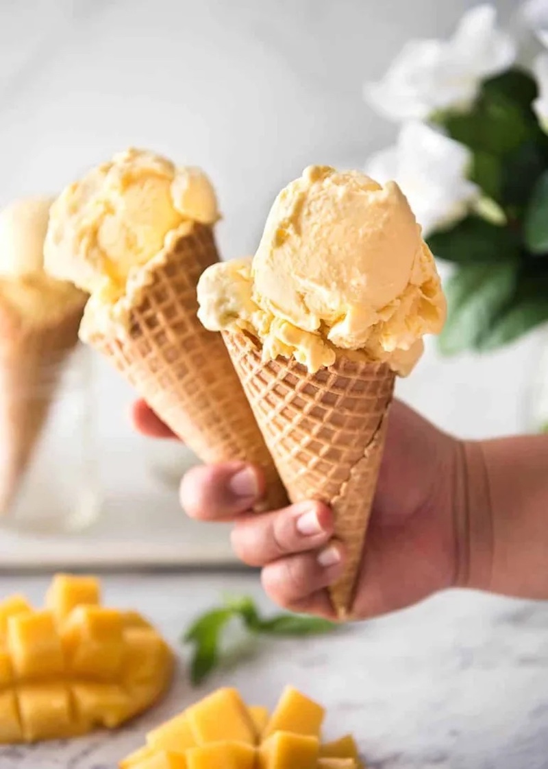 Three waffle cones filled with creamy homemade no-churn mango ice cream, with fresh diced mango in the foreground.