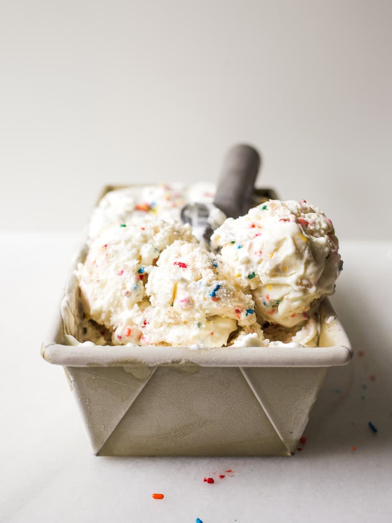 Creamy no-churn sugar cookie ice cream with colorful sprinkles and sugar cookie dough scooped into a loaf pan with an ice cream scoop.