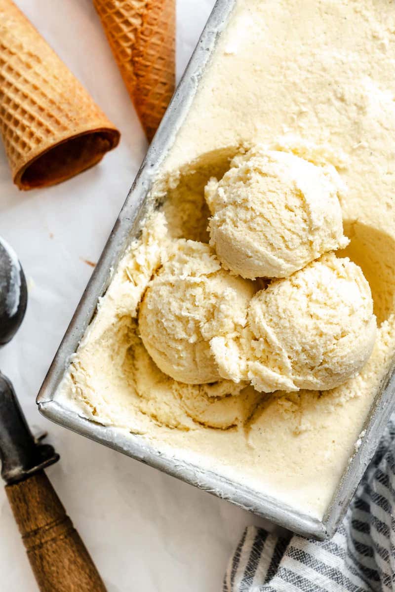 Creamy homemade no-churn vanilla ice cream scooped from a loaf pan with waffle cones nearby.