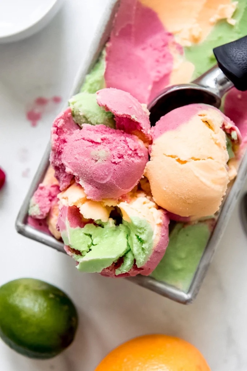Scoops of homemade rainbow sherbet in pink, green, and orange swirls served in a loaf pan with fresh lime and orange nearby.