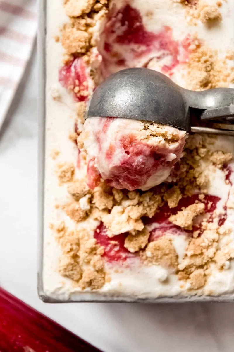 Scoop of roasted rhubarb crumble ice cream with swirls of roasted rhubarb and buttery crumble topping in a loaf pan.