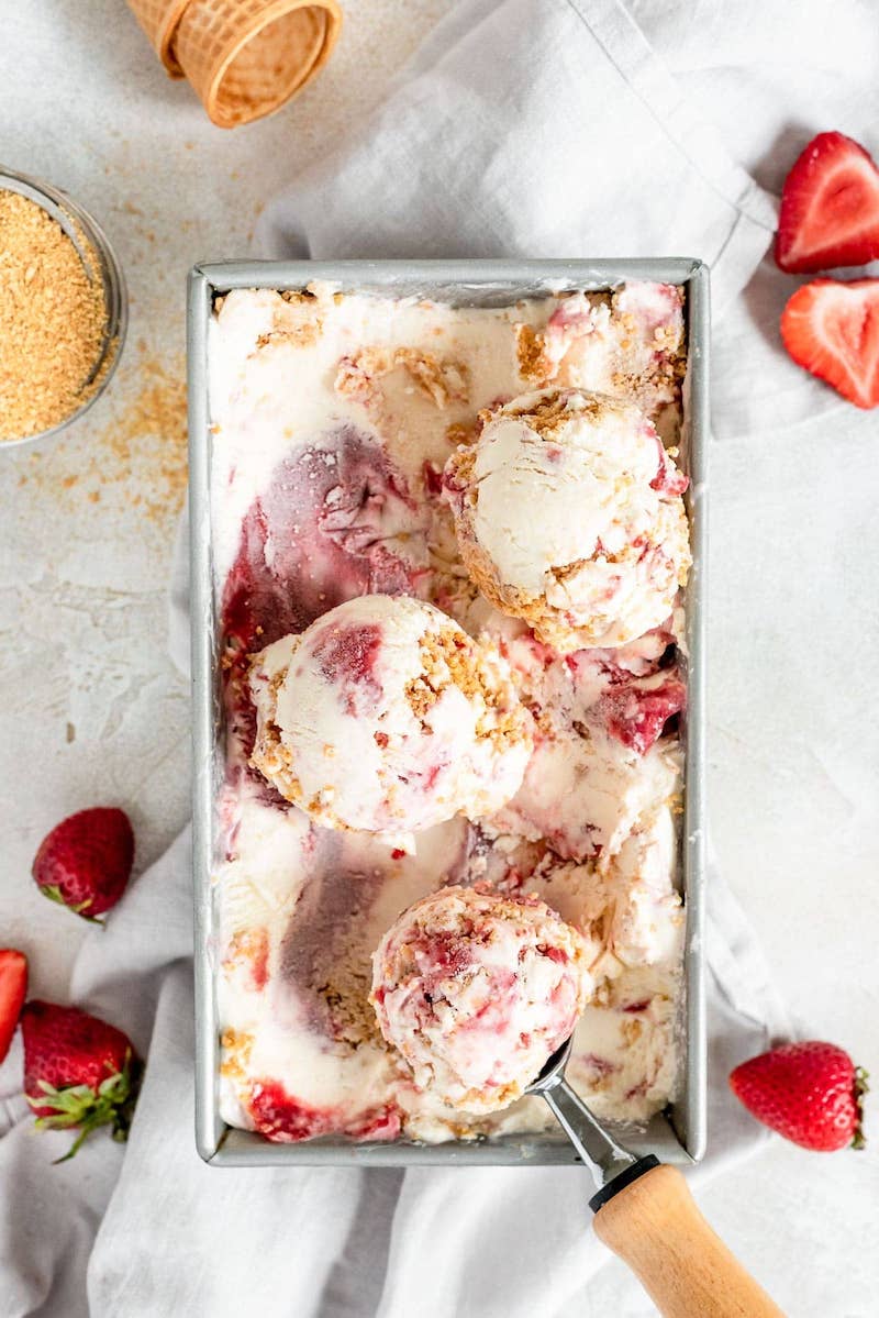 Overhead view of creamy no-churn strawberry cheesecake ice cream swirled with strawberries and graham cracker crumbles in a loaf pan with scoops served alongside fresh strawberries.