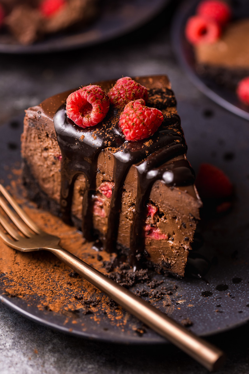 Slice of dark chocolate raspberry cheesecake with fresh raspberries and chocolate sauce on top.